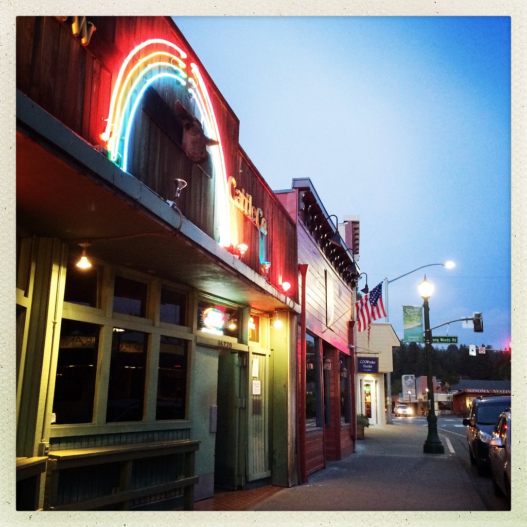The Rainbow Cattle Company at dusk — neon rainbow arching over a steer head on the wooden saloon front, Main Street, Guerneville