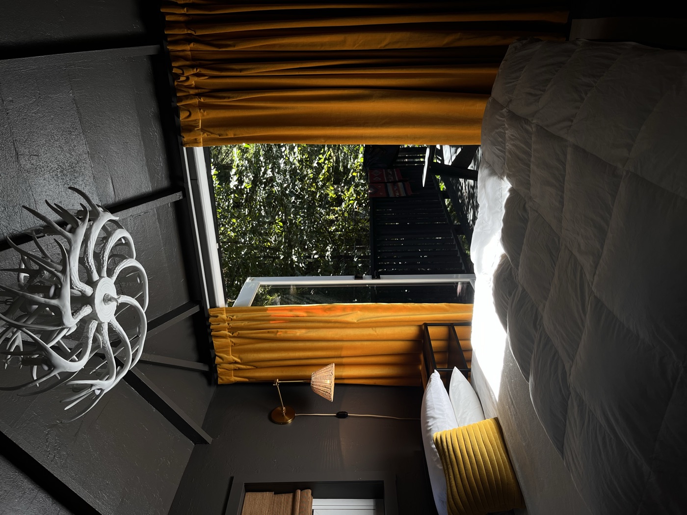 Queen bed beneath a white antler chandelier and charcoal vaulted ceiling, marigold curtains framing french doors open to the redwood deck