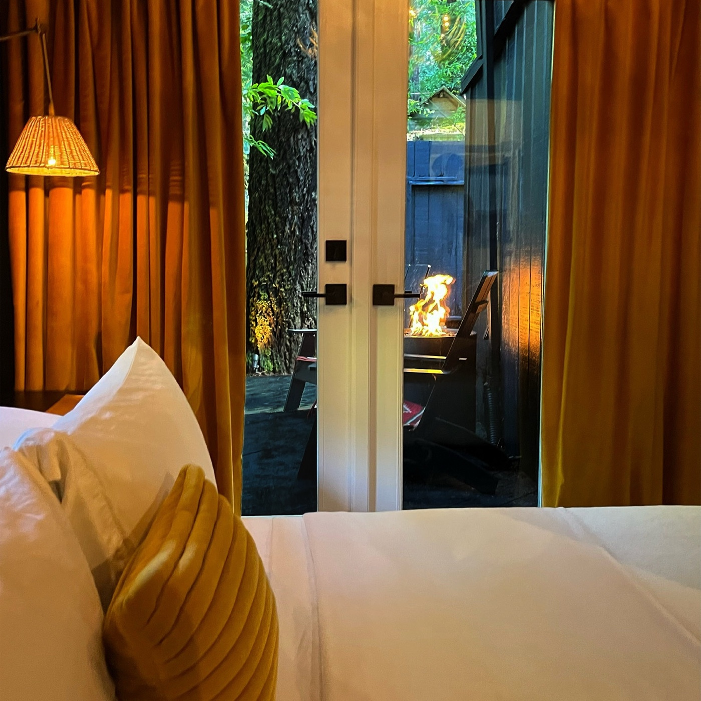 View across the white bed and gold velvet pillow, through french doors to a gas fire table burning on the deck at dusk