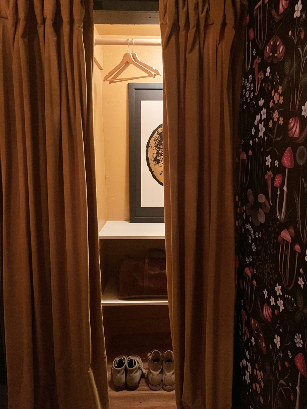 A dressing nook behind mustard velvet curtains — wood hanger, tree-ring art, a leather duffel and boots, botanical mushroom wallpaper alongside