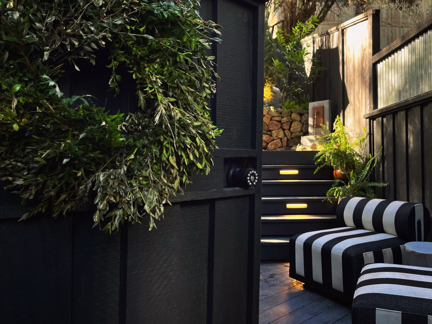 The black board-and-batten cabin at golden hour, a green wreath on the wall, black-and-white striped lounge seating and lit steps rising into the garden