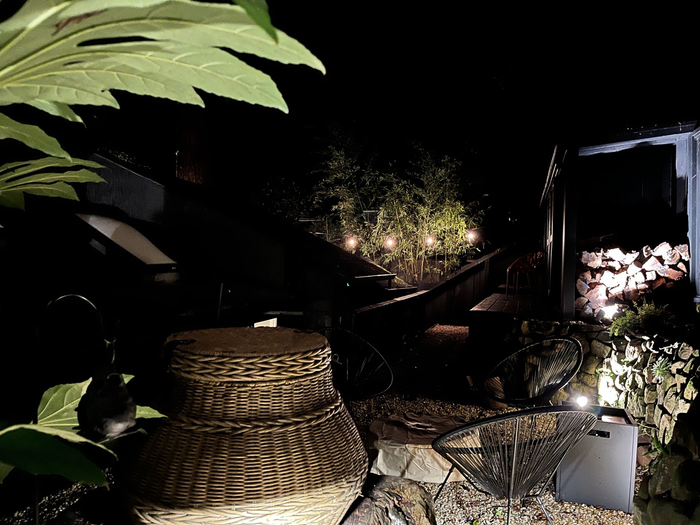 The night patio lit softly — black wire chairs on gravel, a wicker table, a glowing stack of firewood and redwoods rising into the dark