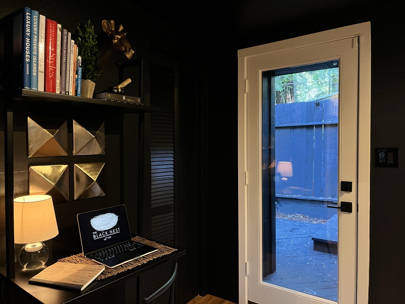 A black desk with design books, a brass giraffe head and gold pyramid art, a laptop showing The Black Nest logo, beside a french door to the deck